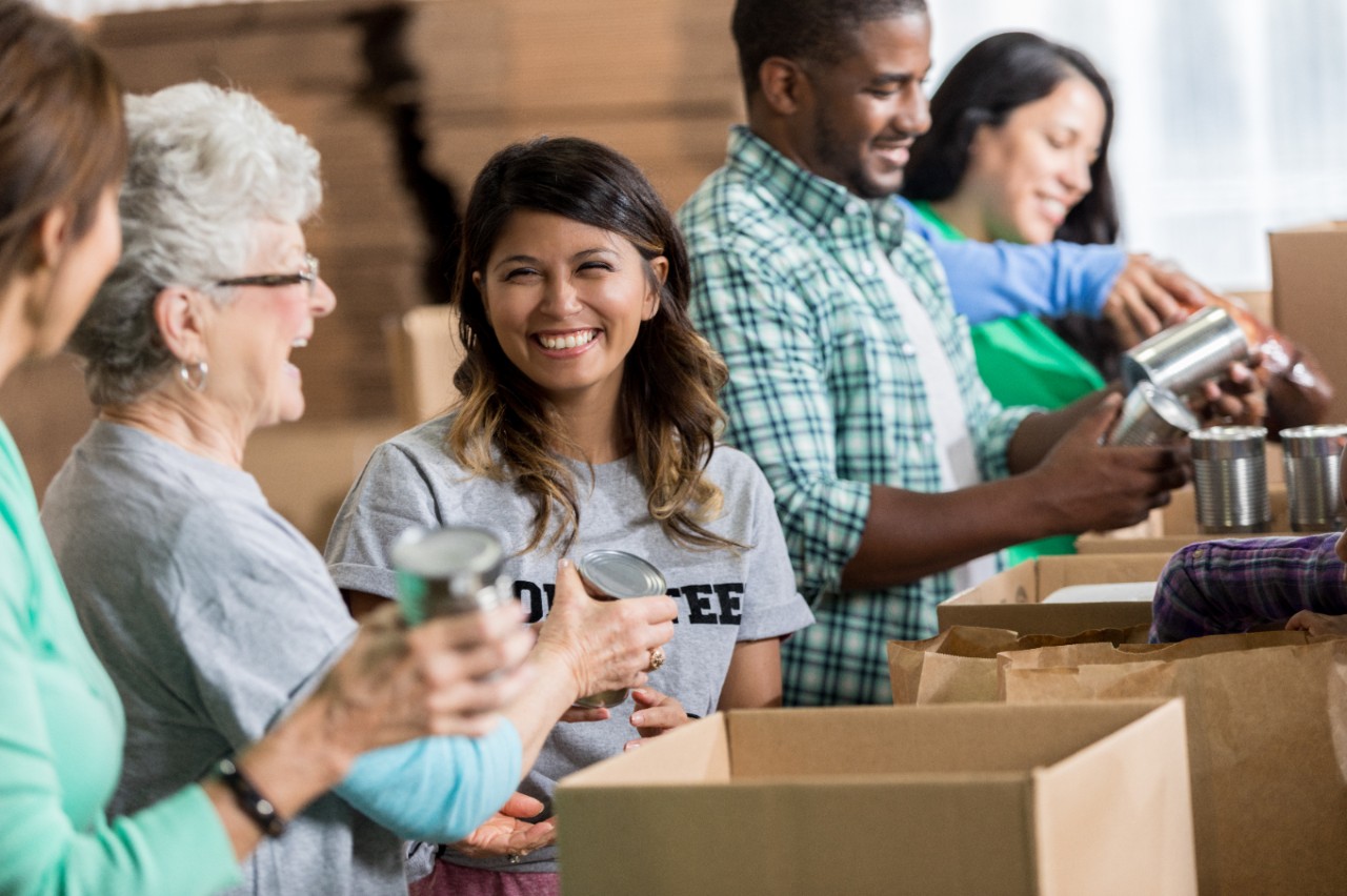 A beautiful Hispanic woman smiles as her senior female Caucasian friend hands her a canned good. They are working in an assembly line while packing food boxes during a community food drive. Other diverse volunteers are working beside them.